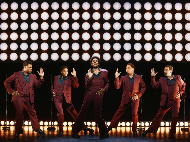 Harrel Holmes Jr., Jalen Harris, Dwayne P. Mitchell, Michael Andreaus, E. Clayton Cornelious from the National Touring Company of Ain’t Too Proud. All are wearing burgundy suits performing a song while dancing. There are many bright lights behind them.