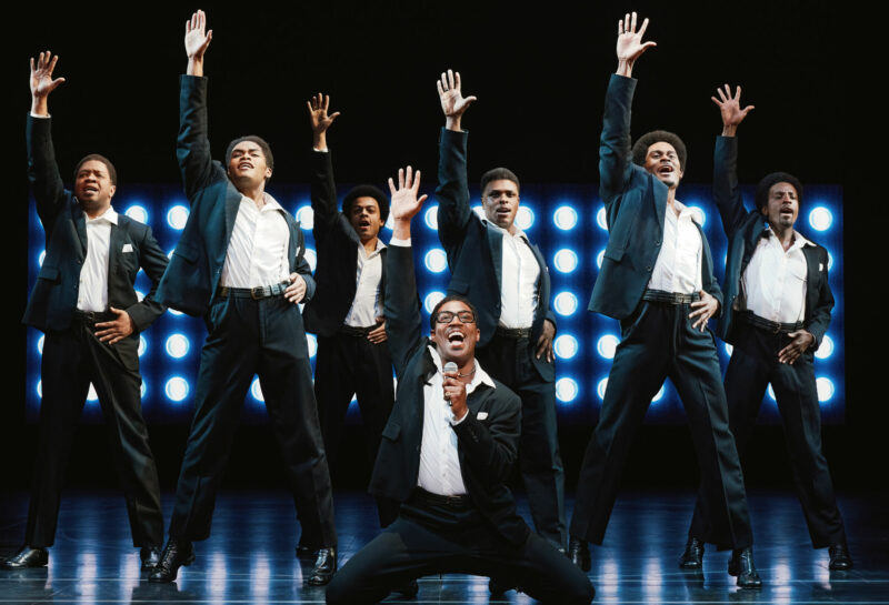 The National Touring Company of Ain’t Too Proud. All are wearing matching black and white suits. They are all performing a song with one arm extended up and there are many bright lights behind them.