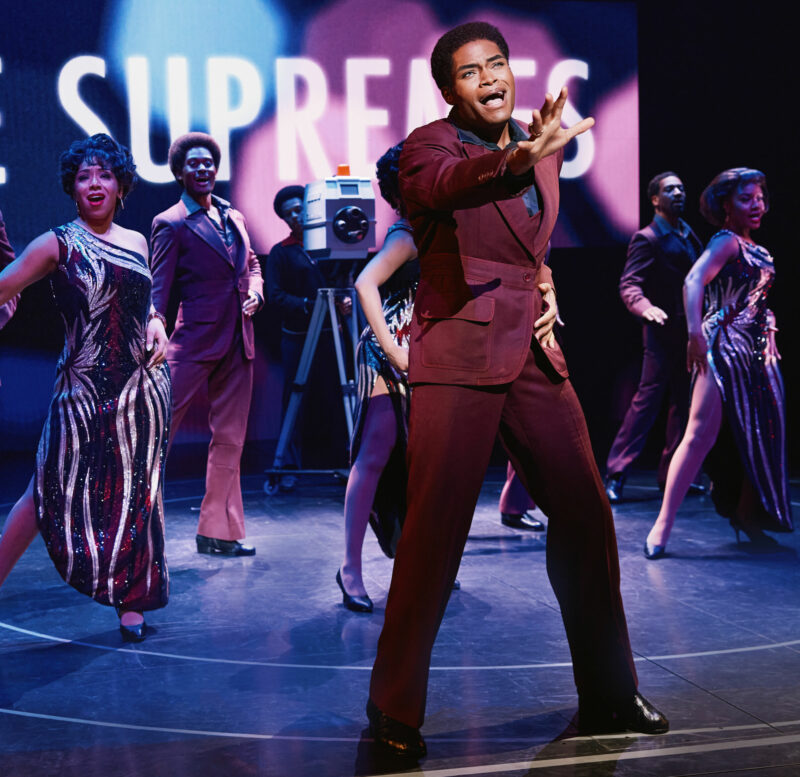 Jalen Harris and the National Touring Company of Ain’t Too Proud. A black man, standing center, is wearing a burgundy suit with one arm stretched out. He is singing while a group of people behind him join in and dance.