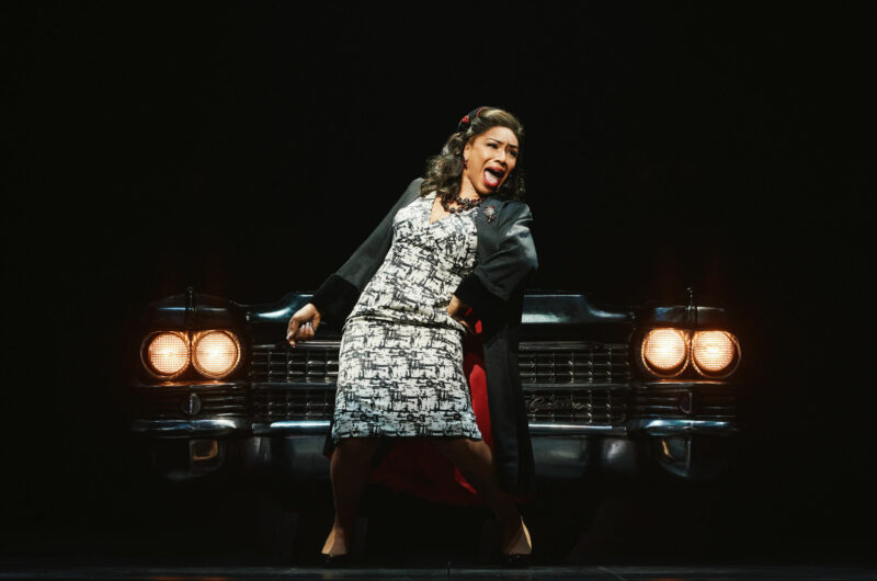 Brittny Smith from the National Touring Company of Ain’t Too Proud. A black woman, wearing a black and white patterned dress, is standing in front of a Cadillac, with its headlights on, performing a song.