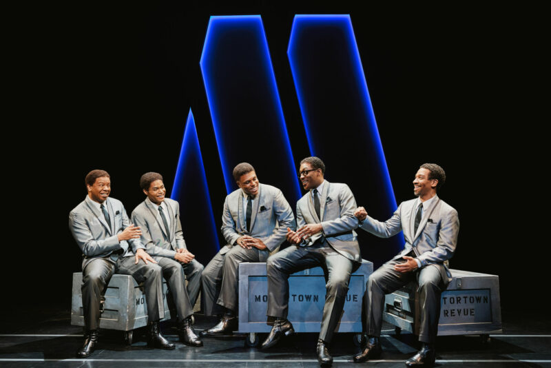 Michael Andreaus, Jalen Harris, Harrell Holmes Jr., Elijah Ahmad Lewis, E. Clayton Cornelious from the National Touring Company of Ain’t Too Proud. All, wearing grey matching suits, are sitting on crates that have “Motortown Revue” on them and a big blue “M” looms behind them.