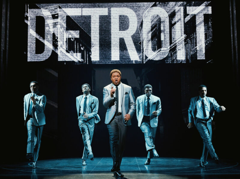 Elijah Ahmad Lewis, Jalen Harris, Michael Andreaus, Harrell Holmes Jr., E. Clayton Cornelious from the National Touring Company of Ain’t Too Proud. All are wearing grey matching suits while performing a song in front of a big sign that reads “Detroit”.